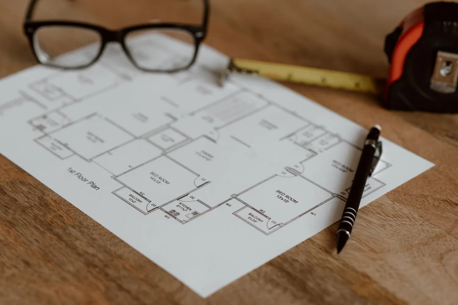 Un plan d'étage imprimé sur un bureau en bois, posé entre un stylo, un mètre ruban et une paire de lunettes