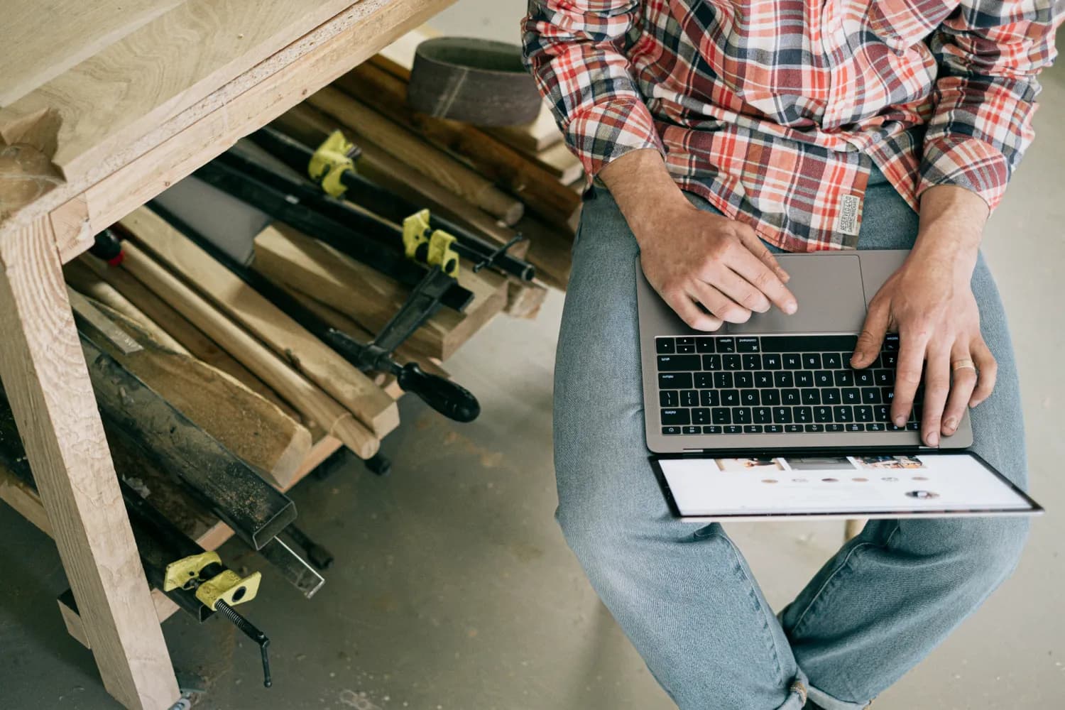 Un menuisier assis sur son établi consulte une boutique en ligne sur son ordinateur portable, à côté d'une pile de planches et de serre-joints