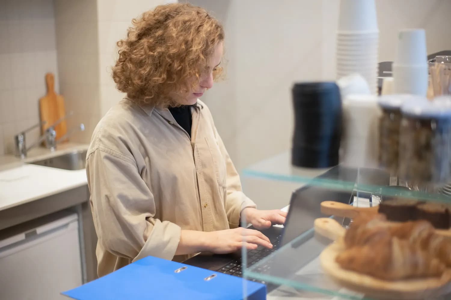 Une commerçante travaille sur son ordinateur portable derrière le comptoir de son café, à côté d'un classeur bleu et d'une vitrine à viennoiseries