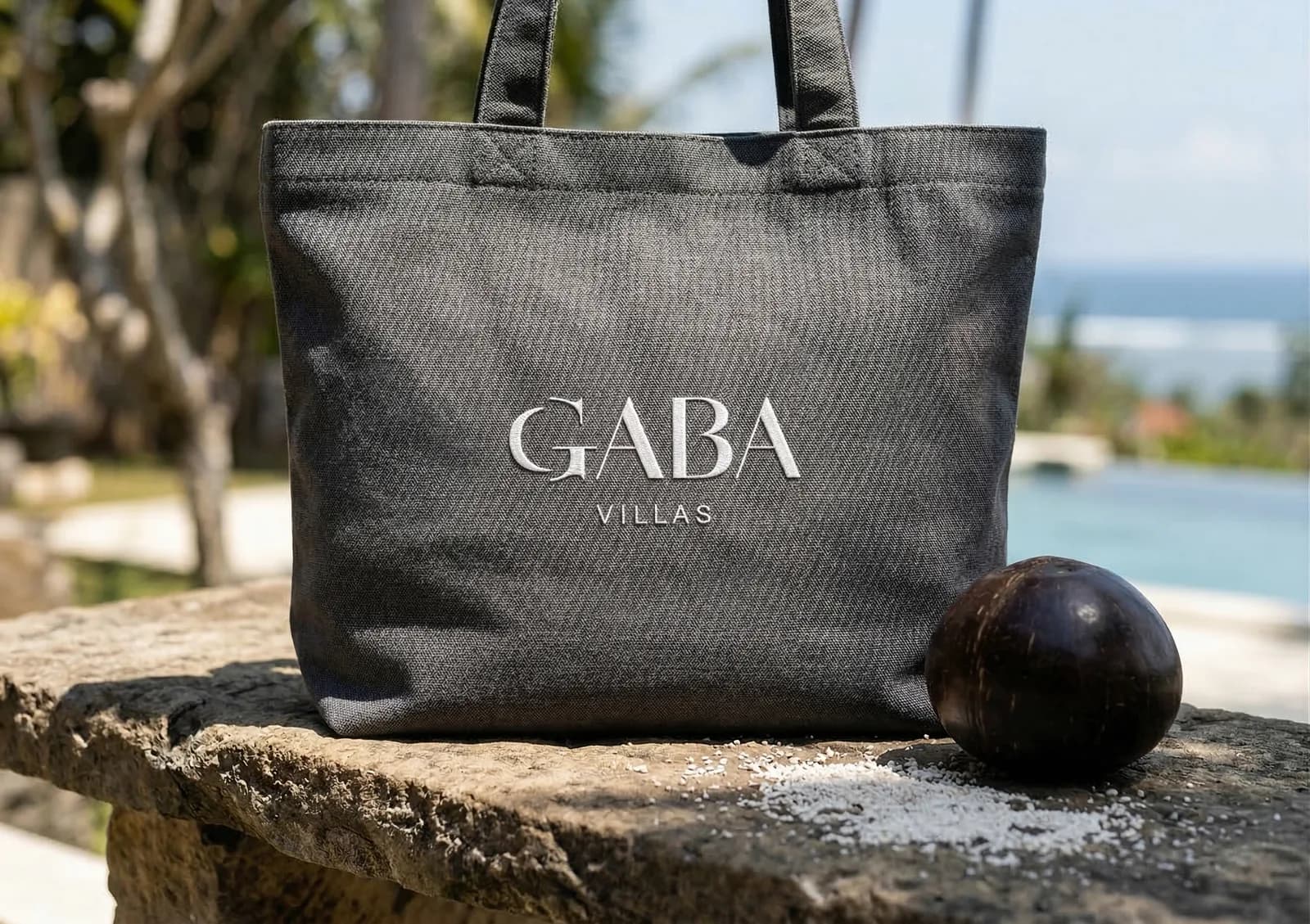 Tote bag en toile grise brodé du logo GABA Villas, posé sur un muret face à la piscine
