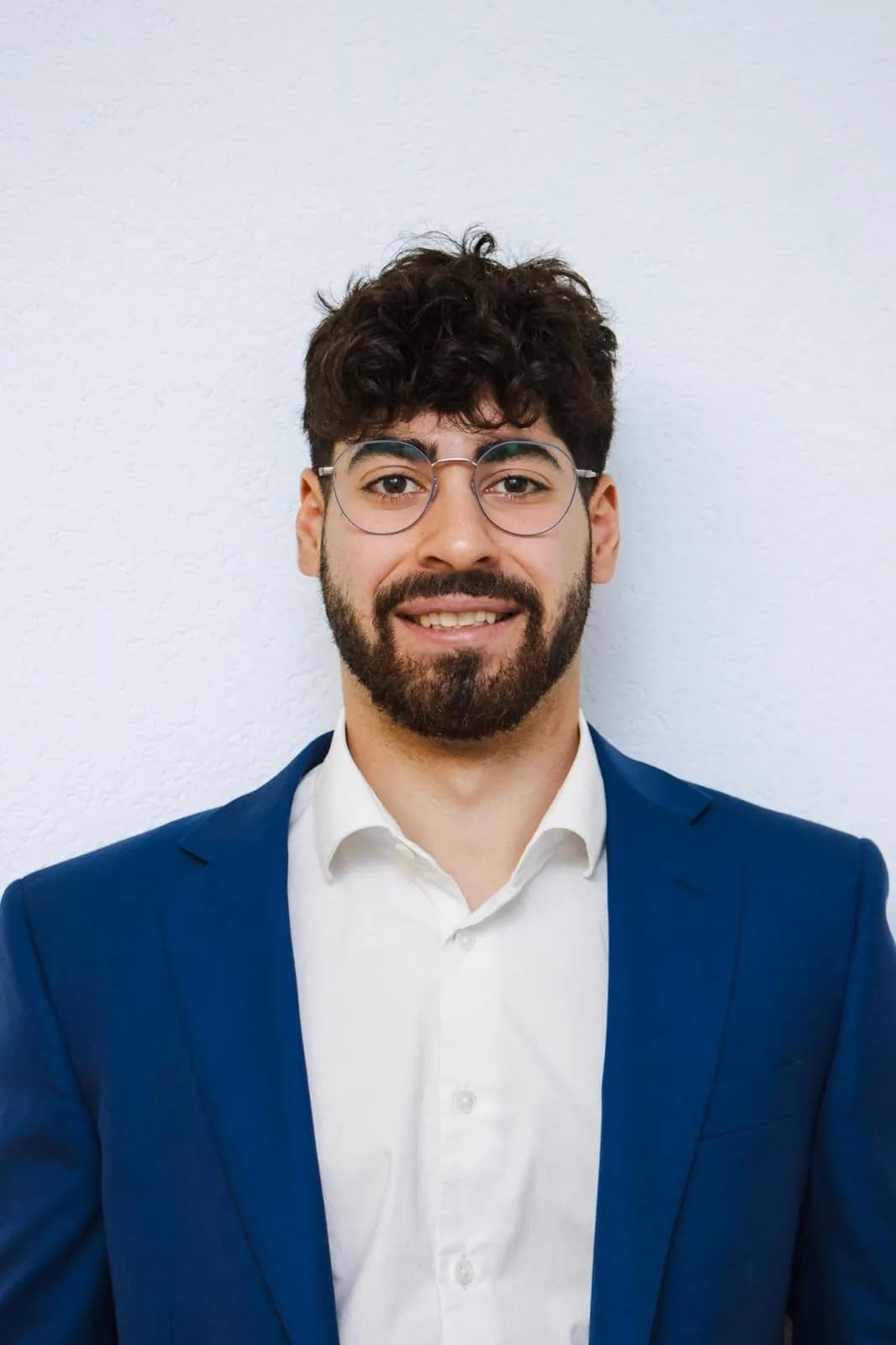 Portrait corporate en buste : veste de costume bleue et lunettes, cadré sur un mur blanc uni.
