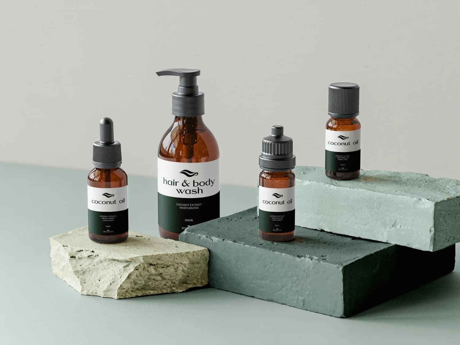 Packshot de quatre flacons ambrés d'une gamme de cosmétiques, posés sur des blocs de pierre vert d'eau, étiquettes blanches et vert forêt lisant « coconut oil » et « hair & body wash »